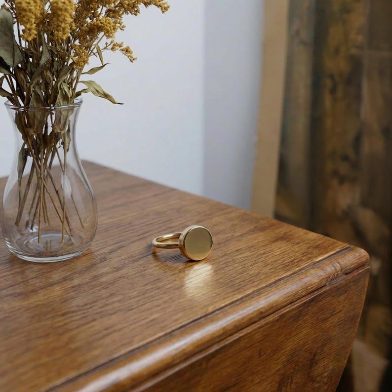 gold round signet ring placed on a wooden surface next to a glass vase with dried yellow flowers, warm natural light, minimal interior setting