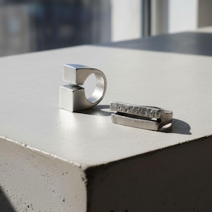 Two modern silver rings displayed on a concrete surface under natural light — one with a clean open-square design, the other a textured geometric bar shape. Industrial minimalist jewelry with sculptural character. Rannka handcrafted pieces.