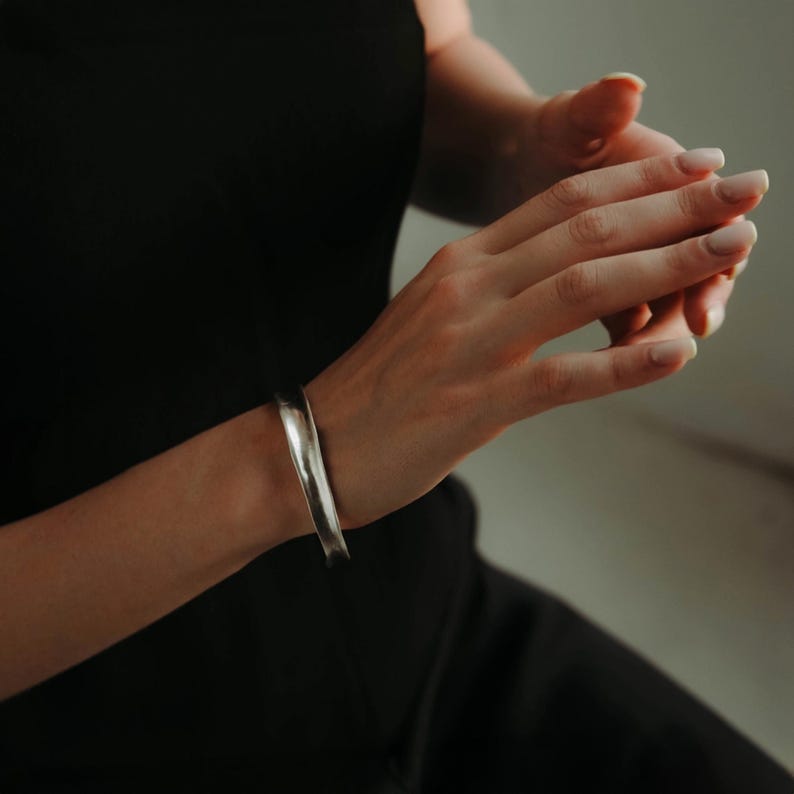 May include: A silver-colored cuff bracelet with a slightly textured surface. The bracelet is worn on a wrist, and the image shows the bracelet's curved shape and metallic sheen. The background is a soft, neutral tone, highlighting the jewelry.