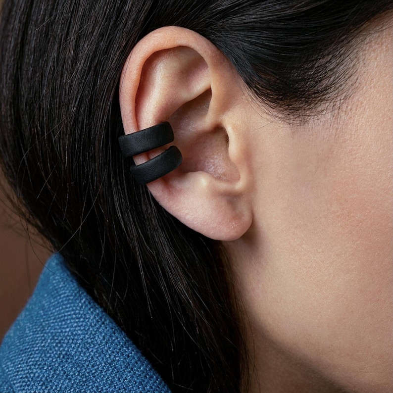 Matte black ear cuffs worn as a duo on the outer ear, no piercing ear accessories hugging the ear edge, minimalist ear wraps with a clean modern look, close-up profile showing stacked placement and smooth curved design by Rannka.