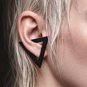 May include: A matte black ear cuff shaped like an open triangle. The geometric accessory wraps around the outer edge of the ear cartilage, featuring clean, sharp lines and a solid, dark finish that contrasts against the skin and hair.