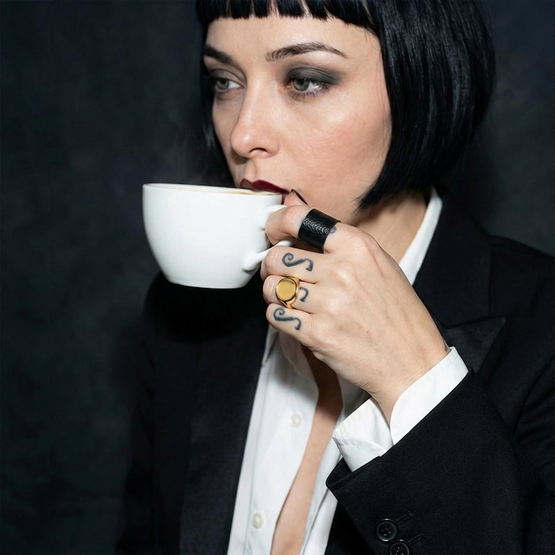 Woman in a black blazer with a short dark bob haircut holding a white cup, wearing a black leather ring and a gold signet ring, with small finger tattoos visible.