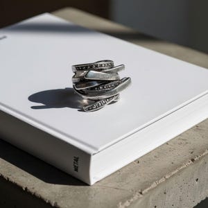 Layered oxidized silver ring with intertwined metal bands and dotted detailing, placed on a white book under natural light. The shadows emphasize its sculptural shape and depth. Handcrafted contemporary jewelry by Rannka with industrial minimalist appeal.