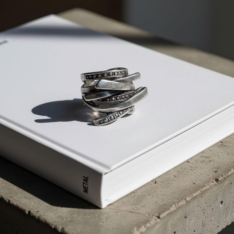 Layered oxidized silver ring with intertwined metal bands and dotted detailing, placed on a white book under natural light. The shadows emphasize its sculptural shape and depth. Handcrafted contemporary jewelry by Rannka with industrial minimalist appeal.
