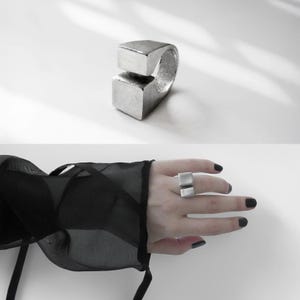 Silver geometric ring with bold square cut design, shown in close-up on a white surface and worn on hand with sheer black sleeve. Contemporary Rannka jewelry, minimalist architectural style with brutalist edge, sculptural modern statement ring.