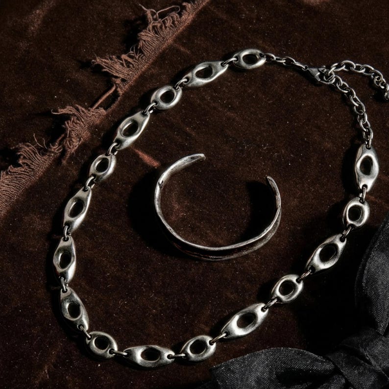 Flat lay of a sculptural silver oval-link chain necklace paired with a minimal open silver cuff bracelet, arranged on rich brown textured fabric with visible weave and frayed edges. Soft directional light highlights the metal patina and organic shapes, creating a moody, tactile, editorial composition aligned with Rannka’s architectural, restrained aesthetic.