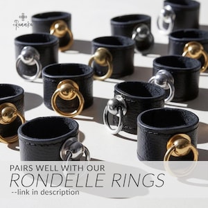 May include: Black leather ring holders with gold and silver metal rings. The holders are arranged on a white surface. The text "PAIRS WELL WITH OUR RONDELLE RINGS" is displayed at the bottom of the image.