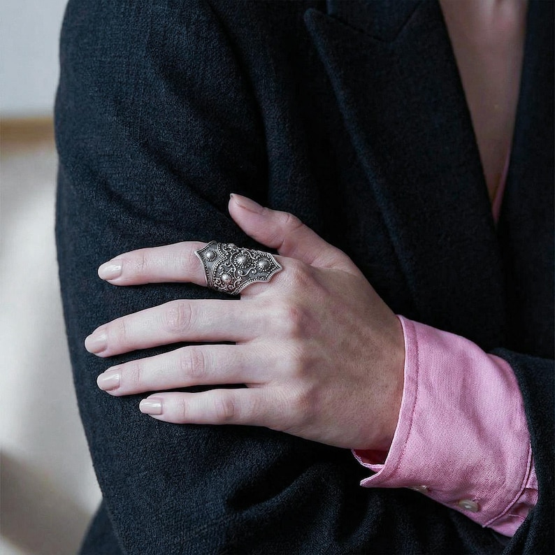 May include: A silver-toned statement ring with an ornate design featuring multiple small, raised circular details. The ring is worn on a hand, with a dark jacket and a pink shirt sleeve in the background.