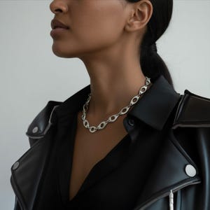 Close-up of a woman wearing a sculptural silver chain necklace with hollow oval links, styled with a black leather jacket and shirt. The look blends modern edge with refined elegance, emphasizing the necklace’s bold, architectural design.