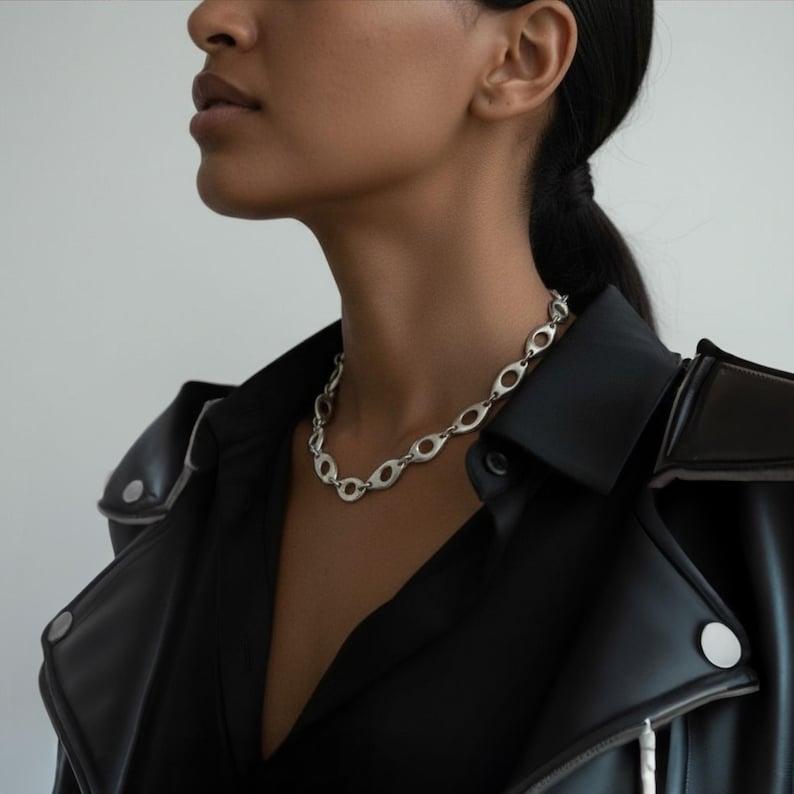 Close-up of a woman wearing a sculptural silver chain necklace with hollow oval links, styled with a black leather jacket and shirt. The look blends modern edge with refined elegance, emphasizing the necklace’s bold, architectural design.