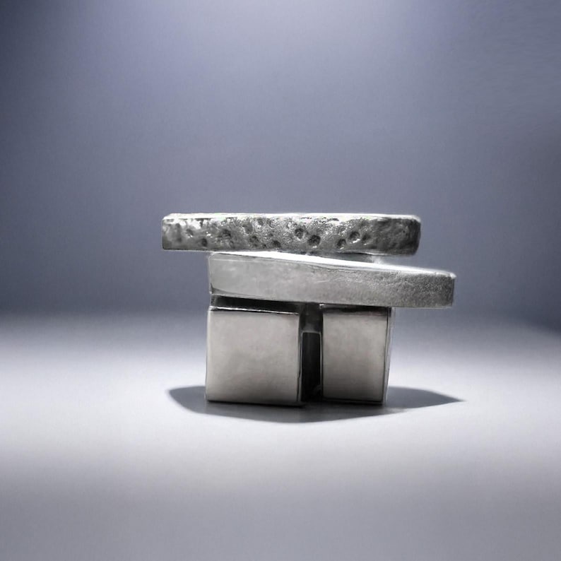 Two silver rings stacked together, one featuring a heavy square cut form and the other layered with elongated hammered bands. Modernist jewelry with raw texture and geometric construction, photographed on a gradient gray background. Rannka design with sculptural, architectural edge and industrial minimalism.