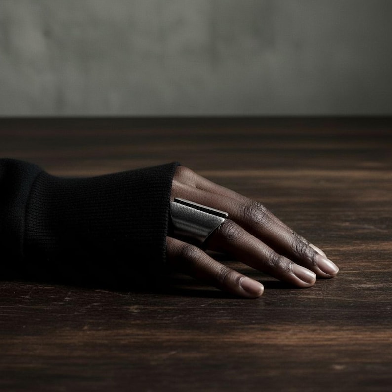 Close-up of a hand resting on a dark wooden surface, wearing a wide silver geometric statement ring. The muted light and black sleeve create a refined, minimalist atmosphere.
