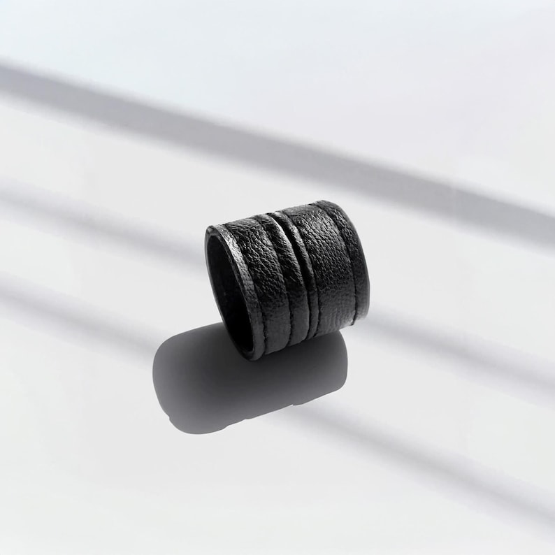 Black vegan leather ring cuff photographed on a clean white surface with soft directional light casting a subtle shadow. Minimal sculptural leather ring with layered wrap detail and matte texture, shown at a slight angle to highlight form and thickness. Contemporary cruelty-free jewelry piece by Rannka, designed as an everyday statement ring with an architectural, understated mood. Suitable for modern styling, gifting, and minimalist wardrobes.