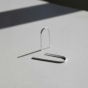 Sterling silver U wire earrings on concrete surface, minimalist open form earrings with smooth rounded top, sculptural arc jewelry design, clean modern silver earrings, simple geometric ear wire, high contrast light and shadow composition, contemporary product styling, Rannka aesthetic