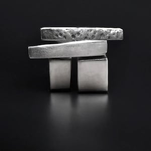 May include: A silver-toned abstract sculpture with a minimalist design. The sculpture features two square blocks supporting a rectangular bar, with a textured bar on top. The piece is set against a dark background.