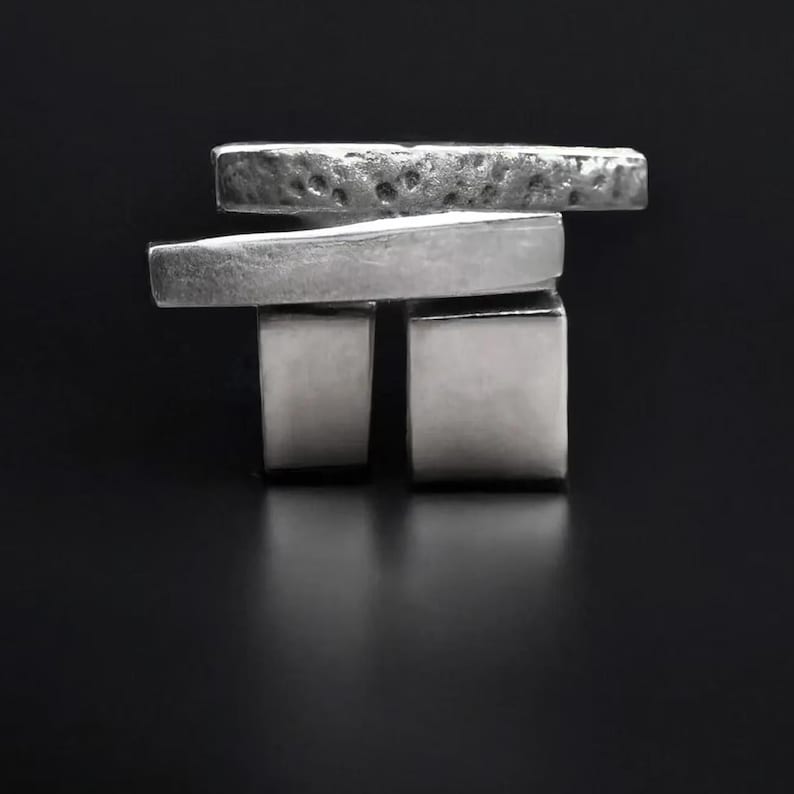 Two brutalist silver rings stacked together, one with bold square block design and the other with layered rectangular hammered bands. Sculptural architectural jewelry by Rannka, photographed on a black background, contemporary industrial aesthetic with geometric forms.