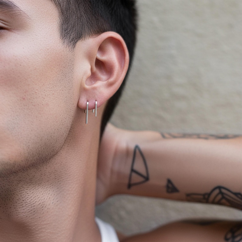 Sterling silver slip earrings in small and medium lengths worn as a clean ear stack, minimalist bar drop earrings on ear, subtle asymmetric styling, modern everyday silver jewelry, close-up ear with short hair and geometric tattoo detail, understated industrial look, Rannka style