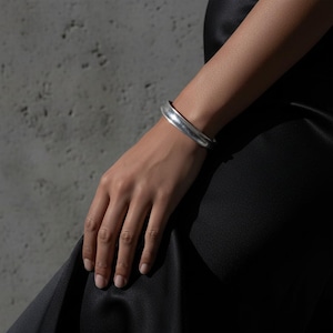 Close-up of a hand wearing a silver cuff bracelet against a concrete wall. The smooth, sculpted band catches natural light, emphasizing its curved form and brushed texture. Styled with a black satin garment for a refined yet industrial contrast, reflecting Rannka’s modern minimal aesthetic.