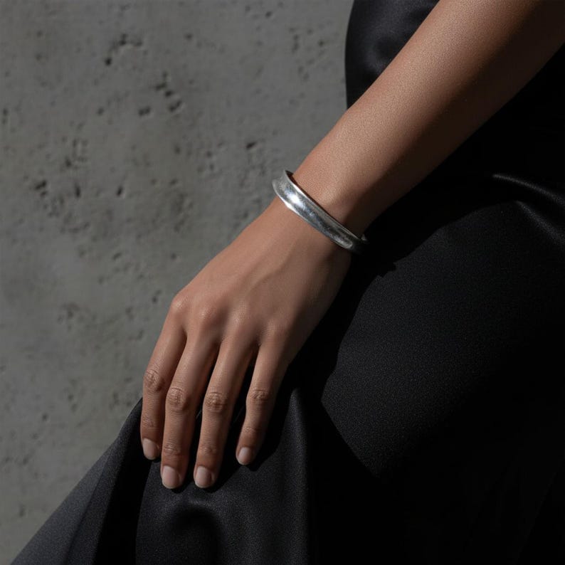 Close-up of a hand wearing a silver cuff bracelet against a concrete wall. The smooth, sculpted band catches natural light, emphasizing its curved form and brushed texture. Styled with a black satin garment for a refined yet industrial contrast, reflecting Rannka’s modern minimal aesthetic.