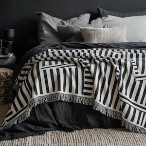 Op de afbeelding: Een zwart-wit patroon deken met franjes, gedrapeerd over een bed. Het geometrische patroon heeft verticale en horizontale strepen. Het bed heeft grijs beddengoed en kussens, met een neutraal gekleurd tapijt op de voorgrond.