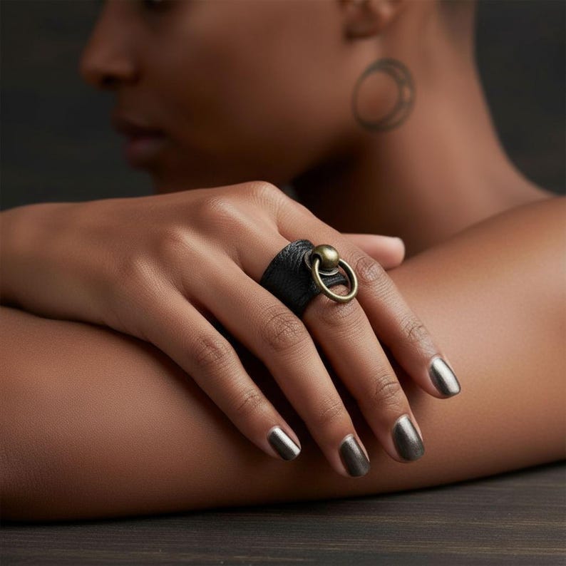 Close-up of hand wearing black vegan leather ring with antique brass sphere and loop accent, resting on arm. Model with metallic nails and geometric tattoo on neck, minimalist dark background highlighting the contrast of textures and sculptural design.