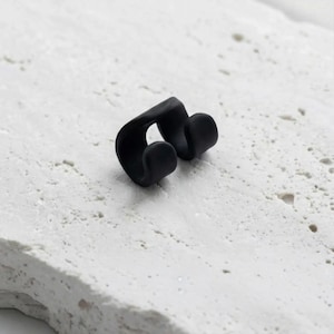 Black double ear cuff resting on a light stone surface, no piercing fake earring with a smooth matte finish. Minimalist adjustable ear cuff designed to wrap the cartilage comfortably. Modern sculptural ear cuff style, lightweight and easy to wear, suitable for everyday use or gifting. Rannka minimalist ear cuff jewelry.