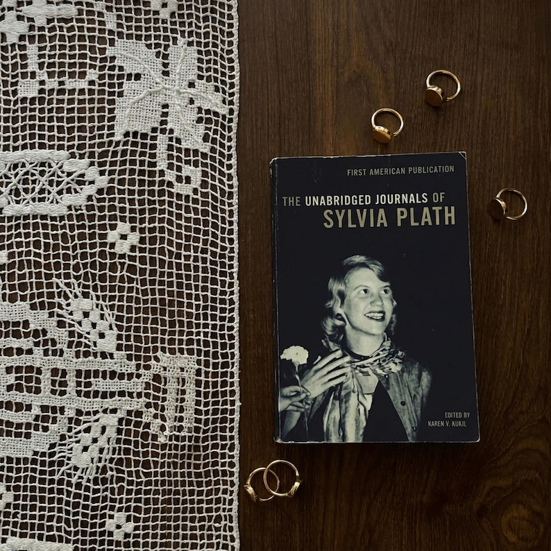 flat lay of a book titled “The Unabridged Journals of Sylvia Plath” on a dark wooden surface, surrounded by multiple 14k gold rings, with a white lace textile placed along the left side of the frame