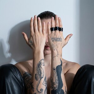 Portrait-style image of a tattooed person seated against a light neutral wall, holding both hands up to partially cover the face. Three black vegan leather wrap rings are worn stacked across adjacent fingers, showing layered band construction and matte texture. Leather pants, and the sculptural ring forms. Contemporary cruelty-free jewelry by Rannka, styled with a raw, modern, architectural mood suitable for statement wear and minimalist gifting.