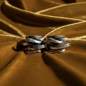 Pair of oxidized silver rings with overlapping layered bands and dotted detailing, placed on a golden fabric background. The metal’s matte and darkened finish contrasts with the soft light reflections of the fabric. Handcrafted statement jewelry by Rannka.