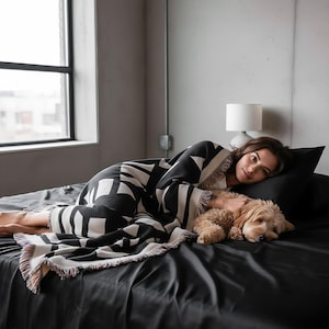 Person lying on a bed with a small dog, wrapped in a black and cream geometric woven blanket with multicolor fringed edges. The scene captures a calm, intimate moment in a minimalist bedroom with concrete walls and soft natural light. Modern textile design by Rannka.