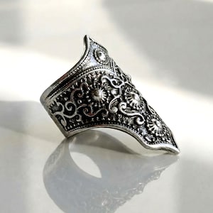 Pode incluir: Anel de armadura de dedo prateado com intrincados desenhos florais e de volutas. O anel apresenta uma forma curva e alongada que cobre parte do dedo. O metal tem um aspeto ligeiramente envelhecido, realçando os padrões em relevo.
