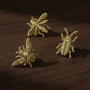 May include: Three gold-colored insect pins are displayed on a dark wooden surface. The pins are intricately detailed, resembling bees and other insects. The pins are made of a woven material, giving them a textured appearance. These pins are suitable for adding a unique touch to clothing or accessories.