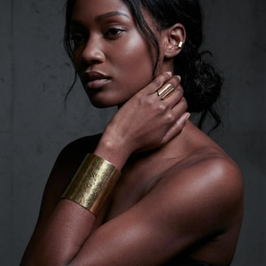 High-fashion portrait of a model wearing a sculptural brushed gold ring, wide gold cuff bracelet, and minimal ear cuff, hand resting at the neck, natural skin texture and soft shadows, editorial jewelry styling with a modern architectural feel, muted tones, studio photography, refined contemporary luxury aesthetic by Rannka.