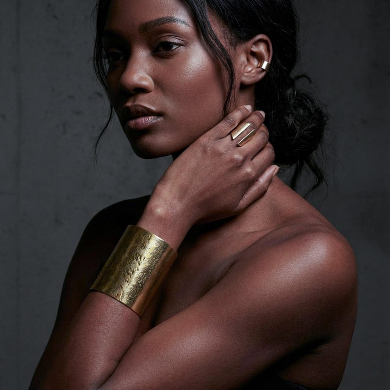 High-fashion portrait of a model wearing a sculptural brushed gold ring, wide gold cuff bracelet, and minimal ear cuff, hand resting at the neck, natural skin texture and soft shadows, editorial jewelry styling with a modern architectural feel, muted tones, studio photography, refined contemporary luxury aesthetic by Rannka.