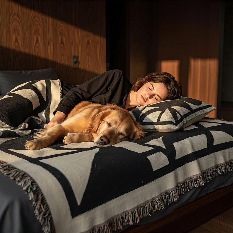 Person sleeping beside a golden retriever on a bed covered with a black and cream geometric woven blanket and matching pillow. The soft evening light and warm wood backdrop create a serene, cozy atmosphere with a minimalist design touch.
