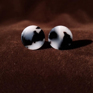 May include: A pair of circular ear plugs featuring a smooth, polished surface with a marbled pattern of black and white swirls. The plugs have a flat, round face and a glossy finish that reflects light, creating a high-contrast appearance.