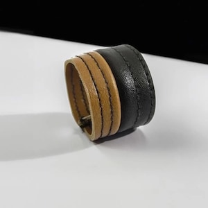 May include: A two-tone leather cuff bracelet. The bracelet features a section of light brown leather with three parallel stitched lines, and a section of black leather. The bracelet is a wide band.