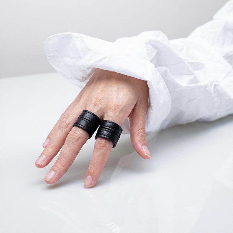 Hand wearing two black vegan leather wrap rings styled across adjacent fingers, photographed on a glossy white surface with soft natural light. Minimal leather ring cuffs with layered band construction and matte texture, shown worn to highlight scale, fit, and finger placement. Clean editorial styling with a white sheer sleeve adds contrast to the dark rings. Contemporary cruelty-free jewelry by Rannka, designed for everyday wear, ring stacking, and modern minimalist gifting.
