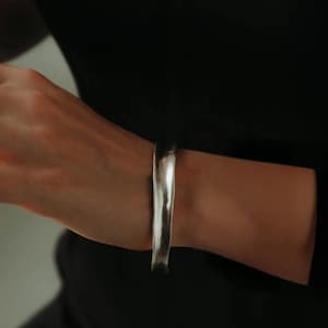 Pode incluir: Uma pulseira de punho prateada com um design texturizado e orgânico. A pulseira é usada no pulso e o metal tem uma superfície polida e reflexiva. A estética geral é minimalista e moderna.