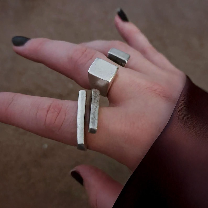 Close-up of hand wearing a brutalist silver ring with stacked geometric blocks and raw industrial texture. Contemporary sculptural jewelry by Rannka, architectural statement design with bold angular forms, photographed outdoors with natural light and dark nail polish for contrast.