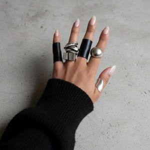 Hand wearing multiple bold contemporary rings, including black band rings, a stacked sculptural metal ring with layered elements, and a smooth rounded statement ring. Mixed textures and architectural forms styled on bare fingers with natural nails. Minimal editorial jewelry styling photographed on a raw concrete background, modern unisex ring collection by Rannka.