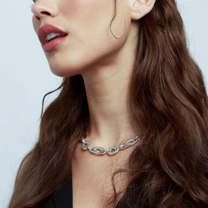 Chunky silver chain necklace worn close to the neck styled as bold statement jewelry with thick hollow links shown on model with long dark hair wearing a black top featuring brushed silver finish and sculptural design often used as heavy chain choker necklace or boyfriend gift for edgy or modern fashion looks