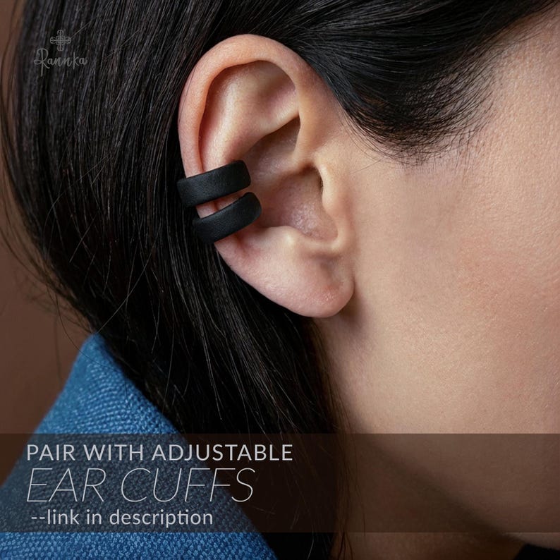 May include: Black adjustable ear cuffs worn on the ear. The ear cuffs are made of a dark material and have a smooth, matte finish. The image also includes the text "PAIR WITH ADJUSTABLE EAR CUFFS".
