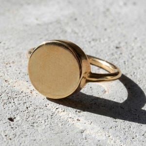 14k gold plated round signet ring, clean flat surface with soft brushed finish, warm light casting a sharp shadow on textured stone background, minimal and solid form with a quiet, refined presence
