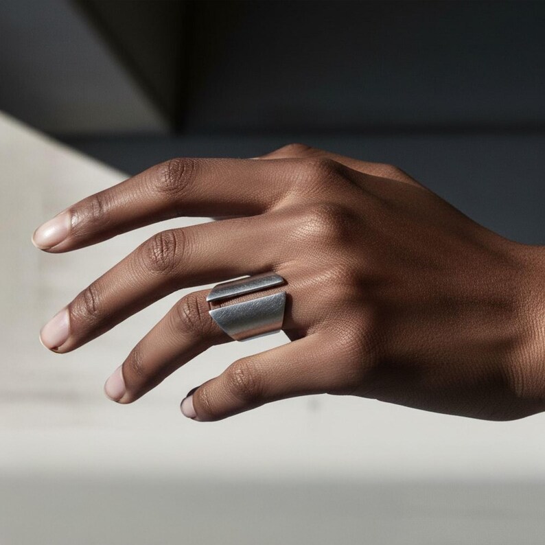 Close-up of a hand wearing a wide silver statement ring with a geometric open-ended design, captured in natural light against a minimalist background.