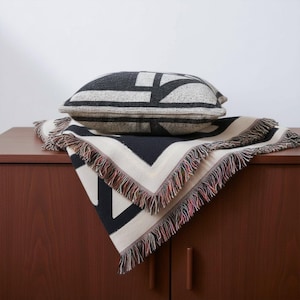 Black and cream geometric woven blanket with multicolor fringed edges displayed with a matching pillow on a wooden cabinet. The bold graphic pattern and neutral tones create a clean, modern look for contemporary interiors.
