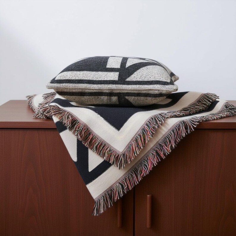 Black and cream geometric woven blanket with multicolor fringed edges displayed with a matching pillow on a wooden cabinet. The bold graphic pattern and neutral tones create a clean, modern look for contemporary interiors.