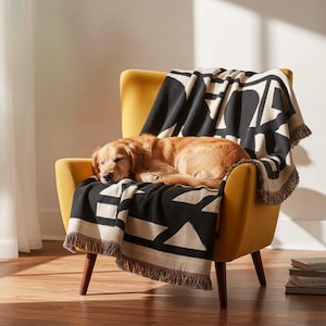 Golden retriever resting on a yellow armchair draped with a black and cream geometric woven blanket featuring multicolor fringed edges. Soft sunlight fills the modern room with warm tones and relaxed comfort.