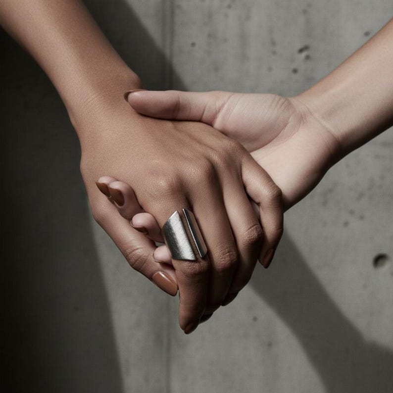 Two hands intertwined against a concrete backdrop, one adorned with a bold silver geometric statement ring — a quiet balance of strength, intimacy, and modern minimalism.