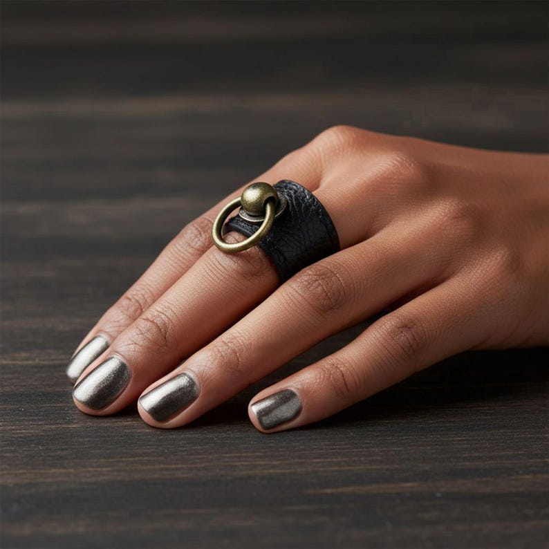 Black vegan leather ring with antique brass sphere and loop detail worn on hand with metallic nails, displayed on dark wood surface. Textured band with bold minimalist hardware accent, modern edgy jewelry design for men or women, photographed close-up to show fit and material.